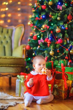 Sevimli bir kız Noel ağacının yanında yerde oturuyor ve Noel süslemeleriyle oynuyor. Yakınlarda bir sürü hediye kutusu var. Tatil sürprizi. Noel zamanı