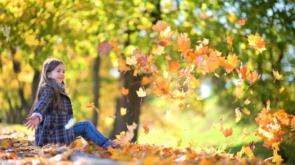可爱的女孩在秋天的公园里玩耍 坐在草坪上扔落叶 — 图库照片