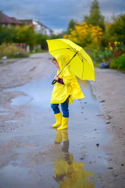 Cute little girl in a raincoat and with a yellow umbrella in her hands walks through puddles, walks in boots and squirts