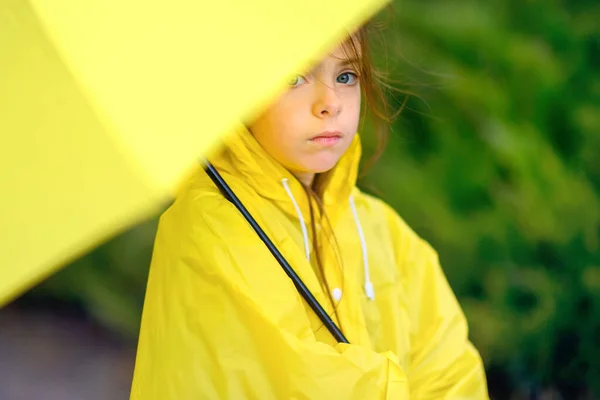 cute little girl in a yellow raincoat is hiding from the rain under an umbrella