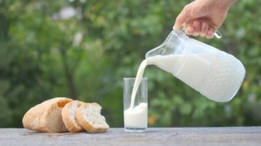 Bir erkek el sürahiden bardağa süt döker. Masanın yanında bir somun ekmek var..
