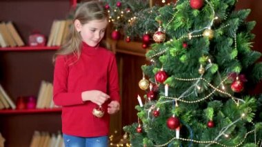 Kırmızı kazaklı tatlı kız odadaki Noel ağacını dekoratif toplarla süslüyor. Noel ve yeni yıla hazırlanıyorum. Tatiller