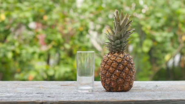 クローズアップの男は 木のテーブルの上のガラスにジュースから新鮮なジュースを注ぎます 近くには天然の緑の背景に熟したジューシーなパイナップルがあります Uhd — ストック動画