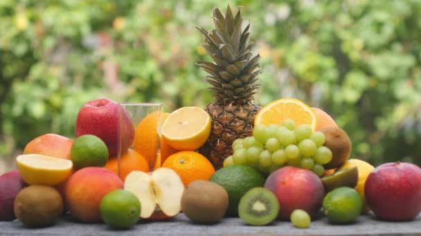 黄色の果実ジュースをガラスに注ぎます 庭の木製のテーブルの上に様々な種類の果物 新鮮なオレンジジュース ソーダのガラス おいしい健康食品 — ストック動画