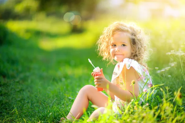Cute girl blows soap bubbles while sitting on the grass in the park at sunset. Children\'s fun.