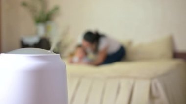 Odadaki havanın iyonlaşmasıyla çalışan bir nemlendiriciden buhar, bulanık bir arka planda, anne bebekle yatakta oynuyor. Nemlendirici işe yarıyor. İklim aygıtı.