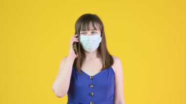 Tıbbi maskeli genç bir kız cep telefonuyla konuşuyor. Sarı arka planda izole edilmiş. ealth bakım ve tıbbi konsept.