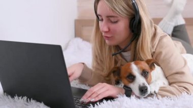 Çevrimiçi iş. Serbest çalışan genç bir kız evinde, yatağında dizüstü bilgisayarıyla çalışıyor. Yanında da sevimli bir köpek yatıyor. Mesafe öğrenme. Ev kavramından çalış. hayvanlar ve insanlar