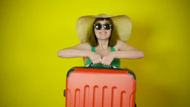 一个戴着大帽子和太阳镜的快乐的年轻女性游客的画像 手里拿着一个手提箱 在一个黄色的工作室背景下微笑着 旅行概念 — 图库视频影像