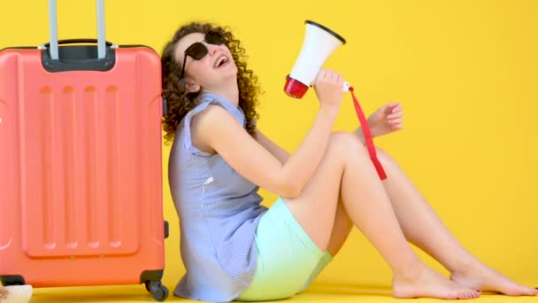 快乐迷人的小女孩 戴着太阳镜 坐在行李箱旁边笑着对着一个被黄色橙色背景隔离的扩音器大喊 航空旅行概念 — 图库视频影像