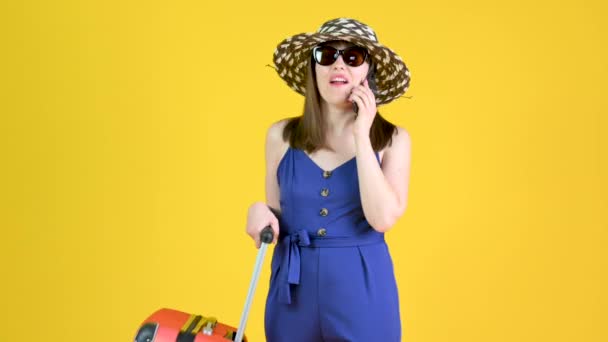 迷人的年轻姑娘戴着帽子和太阳镜 背着手提箱 在工作室里用智能手机把黄色的橙色背景隔开 航空旅行概念 — 图库视频影像