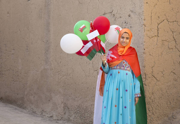 Al Hamra, Oman, November 10th, 2017: Omani girl in national dress with omani flags and baloons 