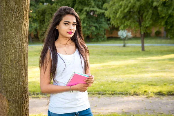 Genç, güzel bir Hintli kız elinde sarı bir defterle bir ağacın yanında duruyor. Dışarıdaki kız öğrencinin portresi.