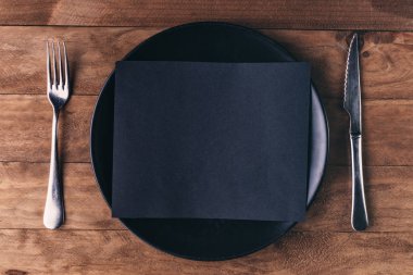 Öğle yemeği davetiyesi, siyah tabak ve ahşap masadaki çatal bıçak takımlarından oluşuyor..