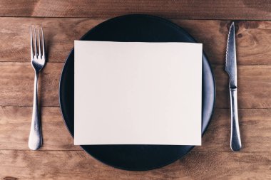 Öğle yemeği davetiyesi, siyah tabak ve ahşap masadaki çatal bıçak takımlarından oluşuyor..
