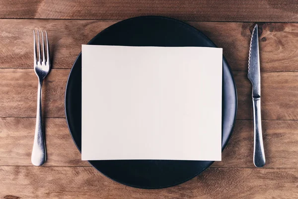 Öğle yemeği davetiyesi, siyah tabak ve ahşap masadaki çatal bıçak takımlarından oluşuyor..
