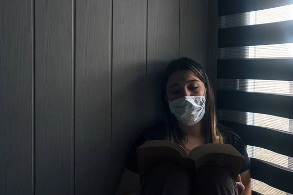Evde karantinaya alınmış bir genç. Pencere kenarında yüz maskesi takmış bir kitap okuyor..