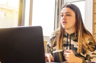 Genç kız terasta ders çalışıyor, dizüstü bilgisayarını kullanıyor ve siyah bardaktan su içiyor..