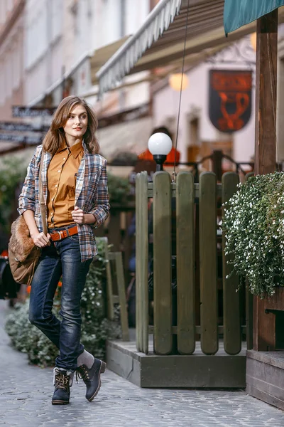 Menina da moda está andando na calçada vestindo jeans azuis, jaqueta quadriculada marrom e segurando uma bolsa marrom, cidade urbana — Fotografia de Stock