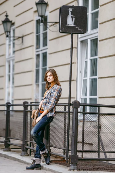 Menina da moda está andando na calçada vestindo jeans azuis, jaqueta quadriculada marrom e segurando uma bolsa marrom, cidade urbana — Fotografia de Stock