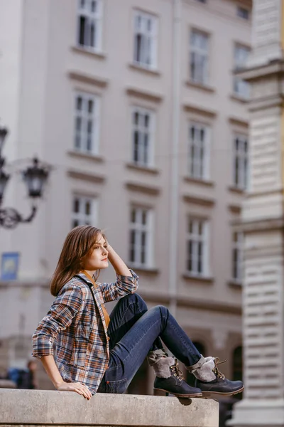 Chica de la moda está sentado al aire libre con pantalones vaqueros azules, chaqueta a cuadros marrón y sosteniendo un bolso marrón, ciudad urbana — Foto de Stock