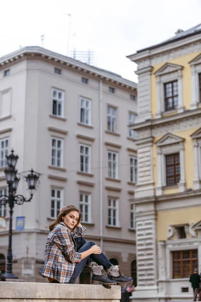 Chica de la moda está sentado al aire libre con pantalones vaqueros azules, chaqueta a cuadros marrón y sosteniendo un bolso marrón, ciudad urbana — Foto de Stock