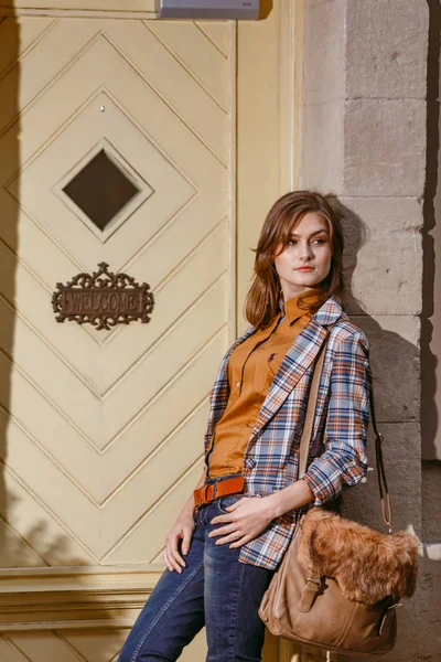 Fashion girl is walking on the sidewalk wearing blue jeans,brown checkered jacket and holding a brown handbag, urban city — Stock Photo, Image