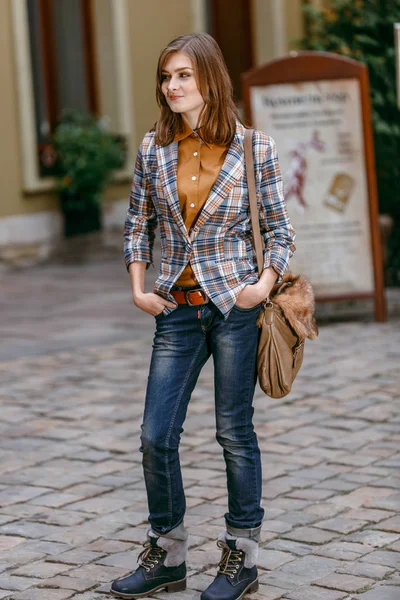 Chica de la moda está caminando en la acera con pantalones vaqueros azules, chaqueta a cuadros marrón y sosteniendo un bolso marrón, ciudad urbana — Foto de Stock