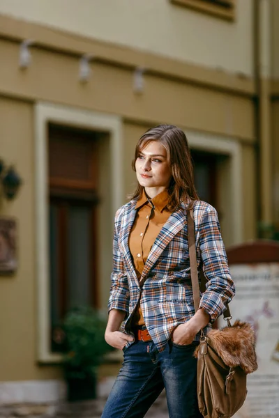 Menina da moda está andando na calçada vestindo jeans azuis, jaqueta quadriculada marrom e segurando uma bolsa marrom, cidade urbana — Fotografia de Stock
