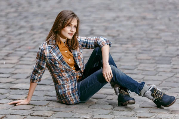 Menina da moda está sentado ao ar livre na rua de paralelepípedos velho vestindo jeans azuis, casaco xadrez marrom e segurando uma bolsa marrom, cidade urbana — Fotografia de Stock