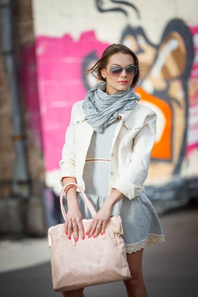 Gyönyörű barna fiatal nő visel, napszemüveget, a könnyű kabát és a kézitáska az utcán — Stock Fotó