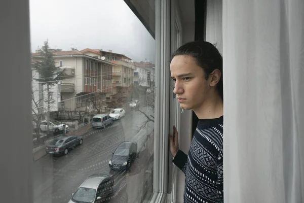 Perdenin önündeki genç endişeli bir şekilde penceresinden boş sokağı izliyor.
