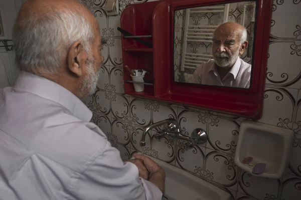 80 yaşındaki Türk adam ellerini yıkarken klasik tuvaletinde aynaya bakıyor.
