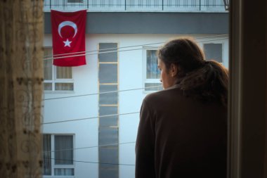 Depresyonda olan, uzun saçlı, balkonundan arka planda Türk bayrağıyla boş sokağı izleyen bir adam.