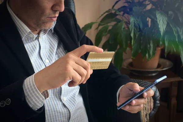 Üst düzey yetişkin erkek şık kıyafetler içinde elinde kredi kartı alışverişi için akıllı telefonunu kullanıyor ve arka planda bir bitkisi var.