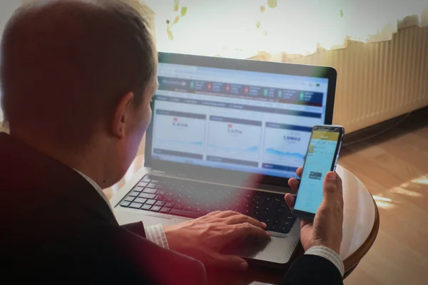 Akıllı giyimli bir işadamı, ev manzaralı bir masada telefon ve dizüstü bilgisayardan borsa analizi yapıyor.