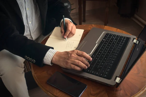 一位专业的老年人一边用钢笔记笔记 一边用笔记本电脑在桌旁打电话 进行电话会议 免版税图库照片