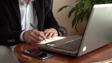 Akıllı profesyonel iş adamı evde dizüstü bilgisayarın başında oturup tele-iş konferansında notlar alıyor.