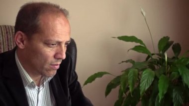 Zeki, üst düzey yetişkin, şık giyinmiş, evde çalışıyor, bilgisayarla maillerini kontrol ediyor ve arka planda bir bitkisi var.