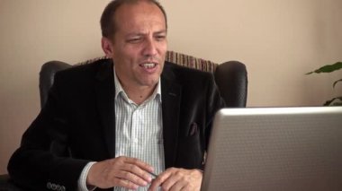 Zeki ve gülümseyen kıdemli iş adamı elinde kalemle evden telgraf çekip iş arkadaşlarıyla laptopuyla görüntülü konuşma yapıyor.