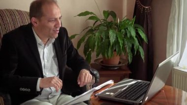 Mutlu zeki ve gülümseyen orta yaşlı iş adamı elinde kalemle evden telgraf çekip iş arkadaşlarıyla laptopuyla görüntülü konuşma yapıyor.