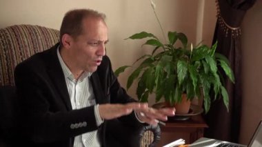 Ciddi görünümlü, orta yaşlı, şık giyinmiş, evinde çalışan ve bilgisayarla yaptığı bir video konferansında iş arkadaşlarına durumu açıklayan bir adam.