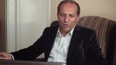Ciddi görünümlü, orta yaşlı, şık giyinen, evde çalışan, bilgisayarını kullanarak iş arkadaşlarıyla konuşan bir adam.