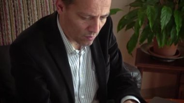 Akıllı bir işadamı evinde oturuyor, günlük giysiler giyiyor, dizüstü bilgisayarı ve telefonuyla borsa alışverişlerini analiz ediyor ve arka planda bir bitkisi var.