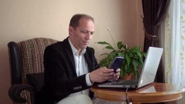 Zeki, mutlu ve gülümseyen, sıradan giysiler içinde dizüstü bilgisayarın önünde oturan ve arka planda bir bitkiyle telefonunu kontrol eden yetişkin.