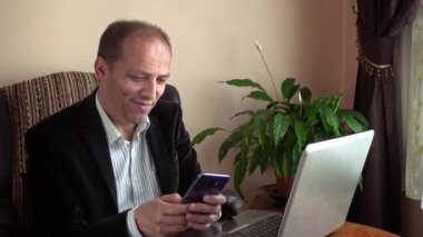 Zeki, mutlu ve gülümseyen son sınıf öğrencisi dizüstü bilgisayarın önünde oturmuş arka planda bir bitkiyle telefonuyla mesajlaşıyor.