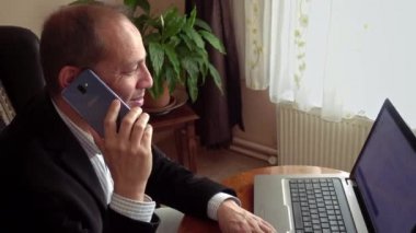 Gülümseyen, orta yaşlı, şık giyinen, evde çalışan, arka planda bir bitkiyle laptopun önünde telefon eden bir adam.