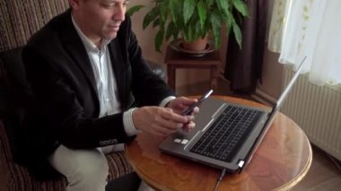 Evde dizüstü bilgisayarla çalışan akıllı, orta yaşlı bir adam bir telefon açar ve arka planda bir bitkiyle konuşmaya başlar.
