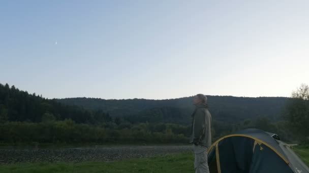 Ein junger Mann steht in der Nähe eines Zeltes, das sich in den Bergen umschaut. — Stockvideo