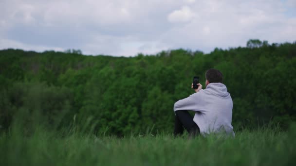 Młody facet odpoczywa robiąc selfie na łonie natury w górach w słoneczny dzień — Wideo stockowe